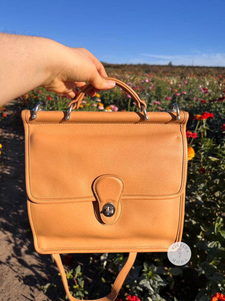 vintage Coach Willis bag in Camel color held in front of a field of flowers. 